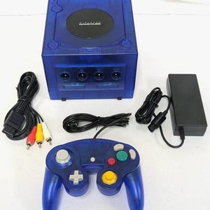 May include: A translucent blue Nintendo GameCube console with a matching blue controller, a black power adapter, and an AV cable with red, yellow, and white connectors. The console features a circular black disc cover with the Nintendo GameCube logo.