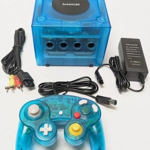 May include: A translucent blue Nintendo GameCube video game console with a matching blue controller, a black power adapter, and a multi-colored AV cable. The console features a circular black disc cover with the Nintendo GameCube logo printed on top.