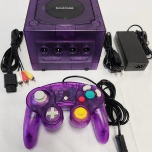 May include: A translucent purple Nintendo GameCube video game console with a matching purple wired controller, a black power adapter, and a multi-colored AV cable. The console features a circular black disc cover with the Nintendo GameCube logo.