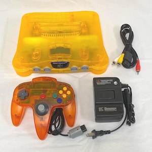 May include: A translucent yellow Nintendo 64 video game console, a matching translucent orange wired controller, a black power adapter, and a black AV cable with yellow, white, and red RCA connectors. The console features the Nintendo 64 logo.