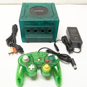 May include: A translucent emerald green Nintendo GameCube console with a matching green controller, a black power adapter, and a multi-colored AV cable. The console features a circular black disc cover with the Nintendo GameCube logo printed on top.