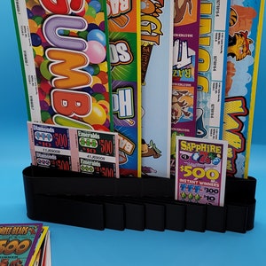 May include: A black lottery ticket holder with multiple lottery tickets in various colors and designs. The tickets include themes such as "Gumballs", "Baseball", "Yoga", "Raz", "Diamonds", "Emeralds", and "Sapphire".