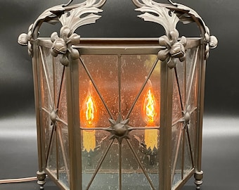 May include: A large, octagonal, bronze lantern with a decorative, floral design. The lantern has a glass panel with a starburst pattern and is lit by two light bulbs.