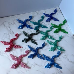 May include: An assortment of colorful dragon figurines in shades of blue, green, pink, and black. The dragon figurines are arranged on a white marble-patterned surface. Each dragon has detailed wings and a textured body.