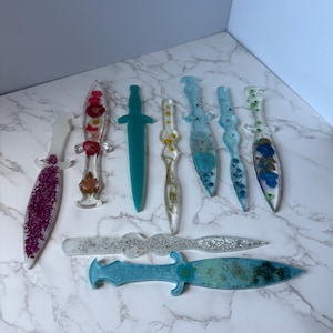 May include: A collection of decorative resin daggers in various colors and designs. Some daggers are clear with embedded flowers, while others feature glitter or solid colors like teal and silver. The daggers vary in size and shape.