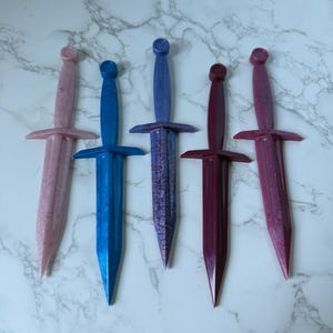 May include: Five decorative daggers in varying colors: pink, blue, purple, burgundy, and reddish-pink. Each dagger has a crossguard and a rounded pommel. The daggers are arranged on a white and gray marble-patterned surface.