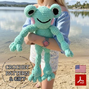 May include: A large, plush, turquoise crochet frog with black eyes and pink cheeks. The frog is being held by a person. The image includes the text "CROCHET PATTERN & GIFT", an American flag, and a PDF icon.