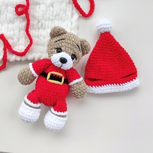 K&ouml;nnte beinhalten: Ein geh&auml;kelter Teddyb&auml;r in einem rot-wei&szlig;en Weihnachtsmannkost&uuml;m mit schwarzem und gelbem G&uuml;rtel, dazu eine passende rot-wei&szlig;e Weihnachtsmannm&uuml;tze und ein wei&szlig;es H&auml;kelst&uuml;ck mit roten Garnakzenten, alles aus weichem, strukturiertem Garn gefertigt.