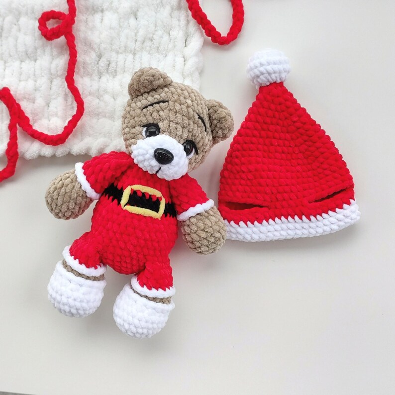 K&ouml;nnte beinhalten: Ein geh&auml;kelter Teddyb&auml;r in einem rot-wei&szlig;en Weihnachtsmannkost&uuml;m mit schwarzem und gelbem G&uuml;rtel, dazu eine passende rot-wei&szlig;e Weihnachtsmannm&uuml;tze und ein wei&szlig;es H&auml;kelst&uuml;ck mit roten Garnakzenten, alles aus weichem, strukturiertem Garn gefertigt.