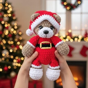 K&ouml;nnte beinhalten: Ein geh&auml;kelter Teddyb&auml;r mit einer rot-wei&szlig;en Weihnachtsm&uuml;tze und einem roten Weihnachtskost&uuml;m mit schwarzem G&uuml;rtel und gelber Schnalle. Der B&auml;r hat hellbraunes Fell, eine wei&szlig;e Schnauze und eine schwarze Nase aus weichem, strukturiertem Garn.