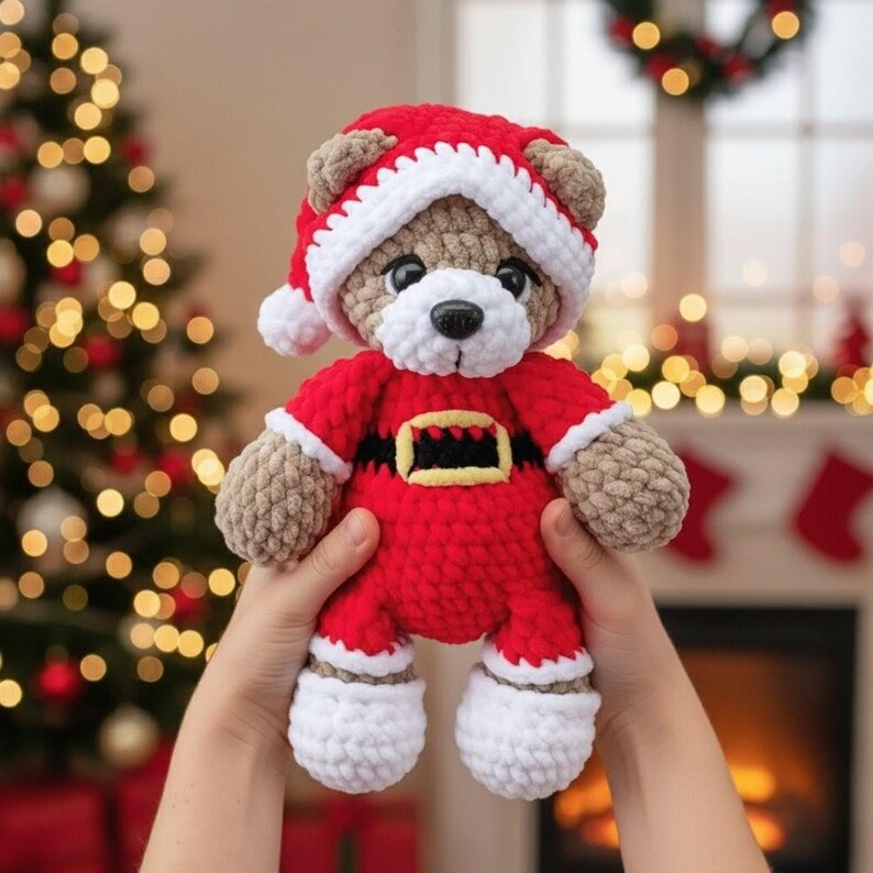K&ouml;nnte beinhalten: Ein geh&auml;kelter Teddyb&auml;r mit einer rot-wei&szlig;en Weihnachtsm&uuml;tze und einem roten Weihnachtskost&uuml;m mit schwarzem G&uuml;rtel und gelber Schnalle. Der B&auml;r hat hellbraunes Fell, eine wei&szlig;e Schnauze und eine schwarze Nase aus weichem, strukturiertem Garn.