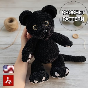 Pode incluir: Um brinquedo de pelúcia de pantera preta de crochê com olhos dourados e detalhes brancos nas patas. A imagem inclui o texto "CROCHET PATTERN" em um design circular. Um ícone PDF e uma bandeira americana também são visíveis.