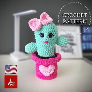 May include: A handmade crochet cactus doll in a pink pot with a heart design. The cactus is teal with a pink bow and smiling face. The image includes the text "CROCHET PATTERN".