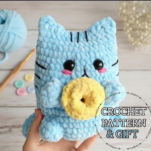 May include: A light blue crochet cat toy with a yellow donut. The cat has black stripes, black eyes, and pink cheeks. The text "CROCHET PATTERN & GIFT" is in a circle on the right. A ball of blue yarn and a crochet hook are in the background.