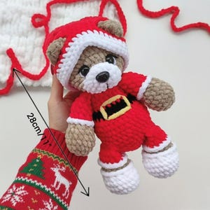 K&ouml;nnte beinhalten: Ein geh&auml;kelter Teddyb&auml;r in einem rot-wei&szlig;en Weihnachtsmannkost&uuml;m mit einem schwarzen und gelben G&uuml;rtel. Der B&auml;r hat braunes Fell, schwarze Augen und eine wei&szlig;e Schnauze. Eine Ma&szlig;angabe zeigt, dass das Spielzeug eine Gesamth&ouml;he von 28 cm hat.