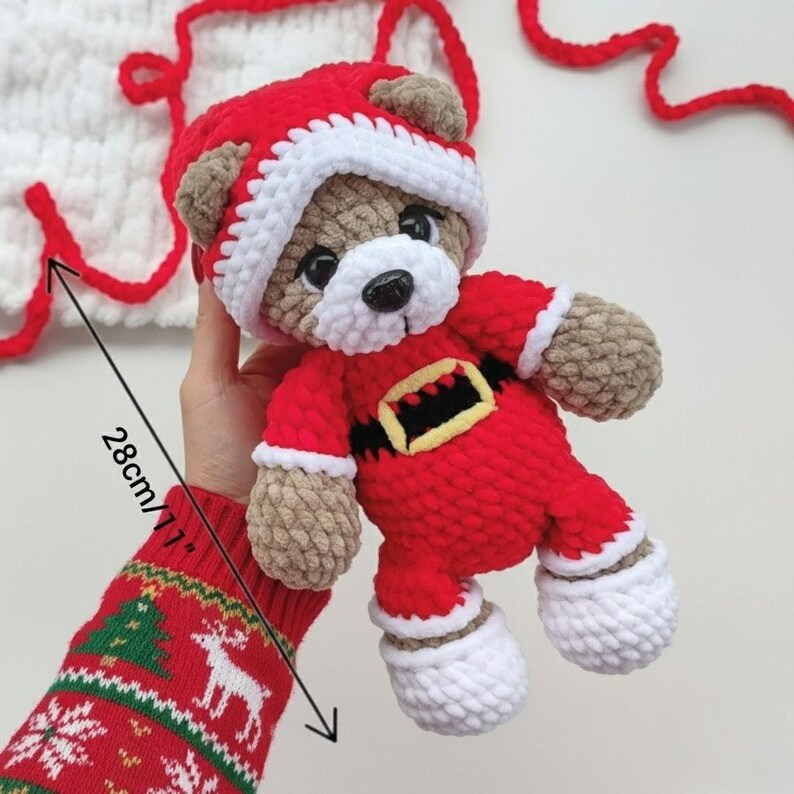 K&ouml;nnte beinhalten: Ein geh&auml;kelter Teddyb&auml;r in einem rot-wei&szlig;en Weihnachtsmannkost&uuml;m mit einem schwarzen und gelben G&uuml;rtel. Der B&auml;r hat braunes Fell, schwarze Augen und eine wei&szlig;e Schnauze. Eine Ma&szlig;angabe zeigt, dass das Spielzeug eine Gesamth&ouml;he von 28 cm hat.