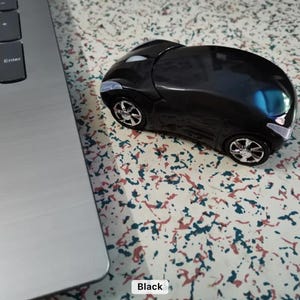 Peut inclure: Une souris d'ordinateur noire en forme de voiture, avec des roues et des phares argentés. La souris est posée sur une surface à motifs, près d'un clavier d'ordinateur portable. Le mot "Black" est dans un encadré blanc en bas de l'image.
