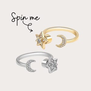 May include: Two open-ended rings, one gold and one silver, each featuring a star and crescent moon design. The star and moon are embellished with small, sparkling stones. The text "Spin me" is written in cursive.