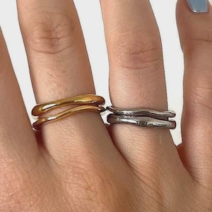 May include: Two stacked rings, one gold-toned and one silver-toned, are shown on fingers. The rings have a fluid, organic design, with a wavy, textured appearance. The gold ring is on the left, and the silver ring is on the right.