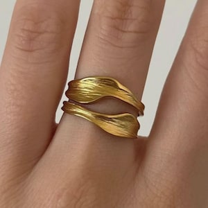 May include: A gold ring with a leaf design. The ring has two overlapping, textured bands, creating a layered effect. The ring is made of a shiny, metallic material.