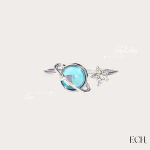 Puede incluir: Anillo plateado para aliviar el estrés con un orbe azul en forma de planeta y un acento en forma de estrella. El anillo tiene un diseño de planeta giratorio. Las palabras "Fidget Ring" y "Spin" están escritas en la imagen.