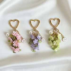 Puede incluir: Tres llaveros florales con cierres dorados en forma de corazón. Cada llavero presenta un racimo de flores en rosa, morado y verde, con una perla y un pequeño dije con la letra "A". Los llaveros están sobre un fondo blanco.