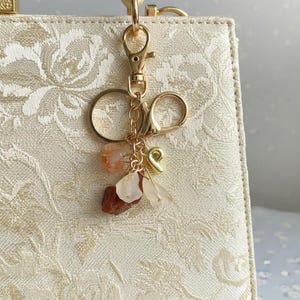 May include: A gold-toned metal keychain featuring a lobster clasp, a circular ring, and a small letter P charm. The keychain is adorned with several irregular, polished stone beads in shades of translucent white, orange, and deep reddish-brown.