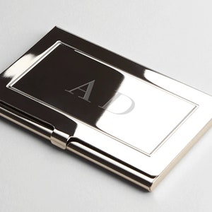 May include: A silver metal business card holder with a rectangular window for displaying a personalized monogram. The monogram is engraved in the window and reads "AD".
