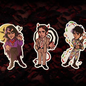 May include: Three illustrated stickers featuring cartoon characters. The first sticker shows a vampire with a purple shirt and cape, the second sticker shows a devil with a red tail and a brown suit, and the third sticker shows a person with a colourful patterned shirt and pants.