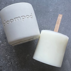May include: Two white ceramic candle holders, one with the word "bloompod" embossed on the side. The candle holder on the right has a wooden stick attached to the top.