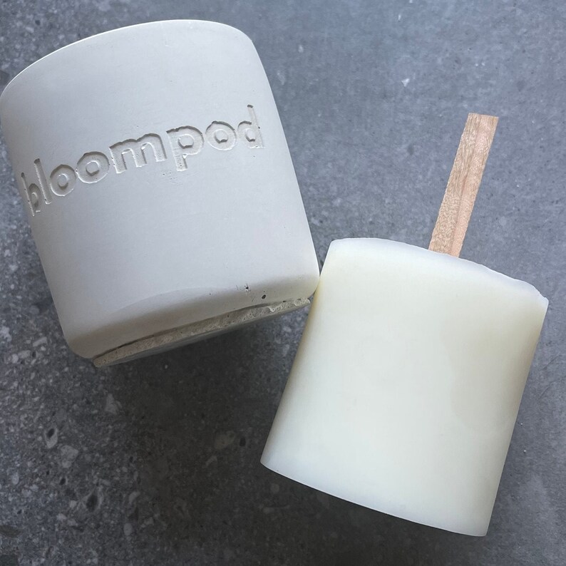 May include: Two white ceramic candle holders, one with the word "bloompod" embossed on the side. The candle holder on the right has a wooden stick attached to the top.