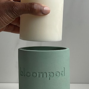 May include: A white candle with a wooden wick sits on top of a sage green concrete candle holder with the word "bloompod" embossed on the side.