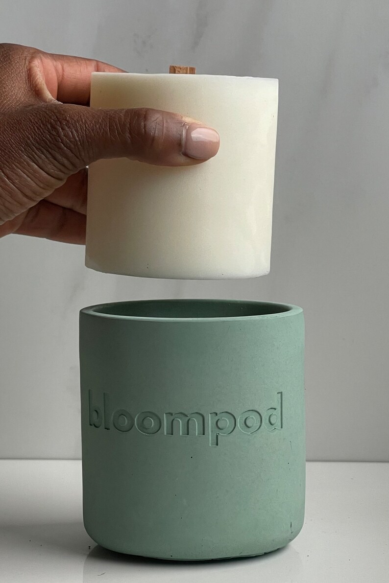 May include: A white candle with a wooden wick sits on top of a sage green concrete candle holder with the word "bloompod" embossed on the side.