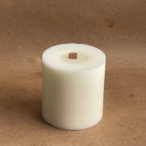 May include: A white pillar candle with a wooden wick. The candle is on a brown surface.