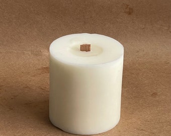 May include: A white pillar candle with a wooden wick. The candle is on a brown surface.