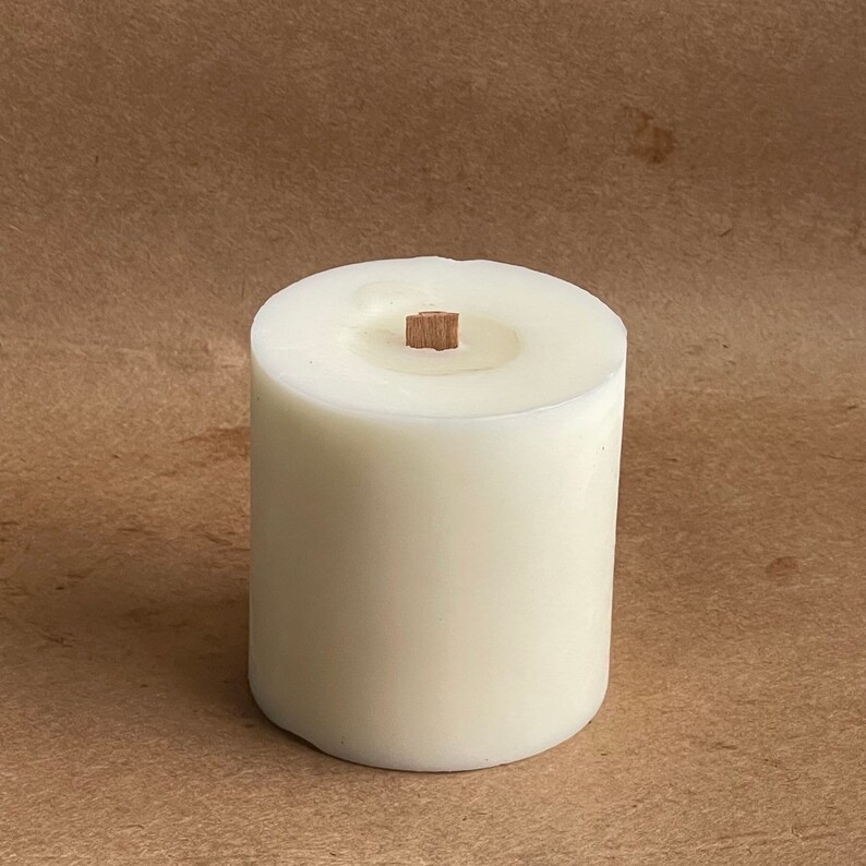 May include: A white pillar candle with a wooden wick. The candle is on a brown surface.