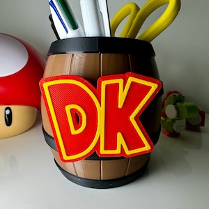 Könnte beinhalten: Ein brauner, fassförmiger Stifthalter mit einem rot-gelben "DK"-Logo auf der Vorderseite. Der Halter enthält Stifte und eine gelbe Schere. Ein roter Pilz und ein grünes Spielzeug befinden sich im Hintergrund.