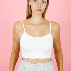 May include: A woman wearing a white cropped tank top and gray sweatpants. She is standing in front of a pink background.