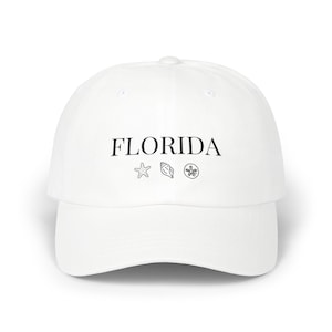 Peut inclure: Casquette de baseball blanche avec le mot "FLORIDA" en noir, accompagné d'un dessin d'étoile de mer, de coquillage et de dollar de sable. La casquette a une visière incurvée et une couronne arrondie.