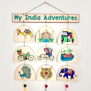 May include: Wooden wall hanging with the text "My India Adventures". It features nine arched wooden panels with colorful illustrations of a camel, Taj Mahal, auto rickshaw, bicycle, hot air balloon, boat, peacock, tiger, and elephant.