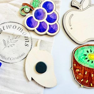 K&ouml;nnte beinhalten: Ein Set aus Holzmagneten in Form von Fr&uuml;chten, darunter eine violette Traubenkette, eine gr&uuml;ne Kiwi, ein roter Apfel und eine gr&uuml;ne Mango. Die Magnete sind handbemalt und tragen das Logo "Just Two Moms Handcrafted Creations".