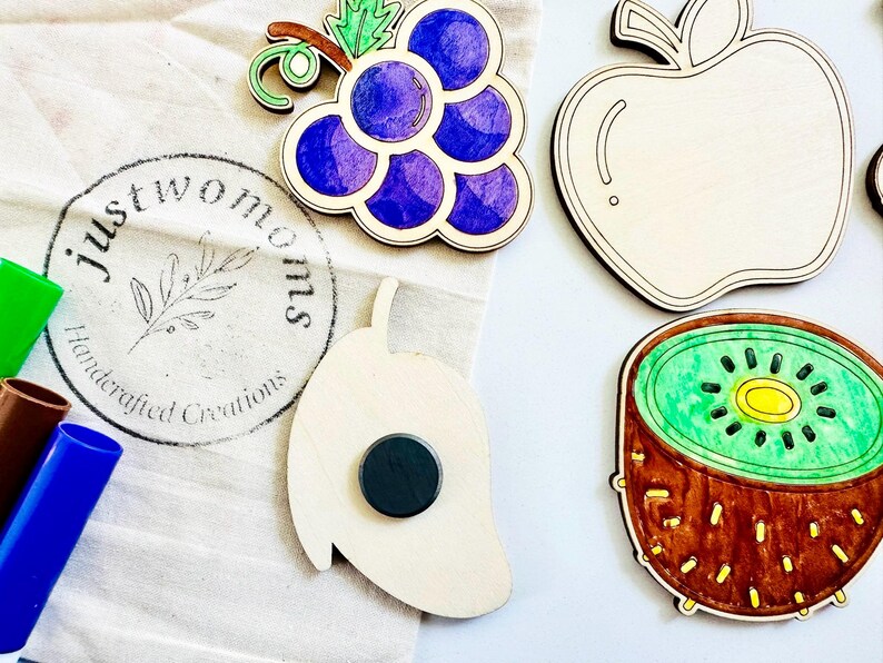 K&ouml;nnte beinhalten: Ein Set aus Holzmagneten in Form von Fr&uuml;chten, darunter eine violette Traubenkette, eine gr&uuml;ne Kiwi, ein roter Apfel und eine gr&uuml;ne Mango. Die Magnete sind handbemalt und tragen das Logo "Just Two Moms Handcrafted Creations".