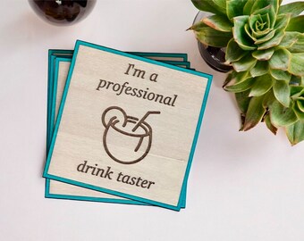May include: Set of four wooden coasters with a teal border. The top coaster features a brown illustration of a coconut drink with a straw and the text "I'm a professional drink taster".