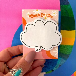 Può includere: Un blocco note adesivo bianco a forma di nuvola con un contorno nero. Il blocco note è su uno sfondo rosa e arancione con margherite bianche e il testo "Ceigh Studios".