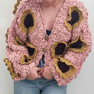 Può includere: Un cardigan lavorato all'uncinetto a maglia grossa in una tonalità rosa chiaro, con scollo a V e due bottoni rotondi marroni. Il capo è decorato con molteplici applicazioni floreali tridimensionali, ciascuna con un centro marrone scuro e petali giallo senape.