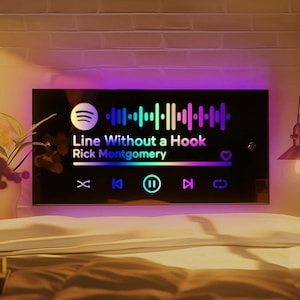 Op de afbeelding: Een zwart rechthoekig bord met een regenboogkleurige Spotify-streepjescode en de tekst "Line Without a Hook Rick Montgomery". Het bord heeft een harticoon en muziekspelerbesturingselementen.