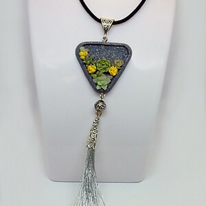 May include: A silver necklace with a grey triangular pendant featuring a succulent garden design. The pendant is attached to a black cord and has a silver tassel.