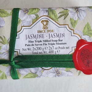 May include: A rectangular soap bar wrapped in floral paper with white jasmine flowers and green leaves. The bar is tied with a green ribbon and sealed with a red wax stamp featuring a heart design. The label reads "JASMINE - JASMIN".