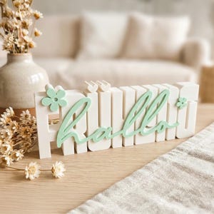 Könnte beinhalten: Ein weißes "Hallo"-Schild in Blockbuchstaben mit mintgrüner Schreibschrift-Überlagerung. Das Schild hat Blumen- und Vogelakzente. Es steht auf einer Holzoberfläche, mit Trockenblumen und einer Vase im Hintergrund.