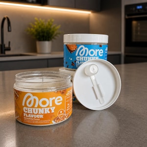 Könnte beinhalten: Drei transparente Gläser des Produkts "More Chunky Flavour", eines mit offenem Deckel und einem Löffel. Die Gläser haben orange und blaue Etiketten mit Produktname und Geschmack. Das offene Glas zeigt das Produkt im Inneren.